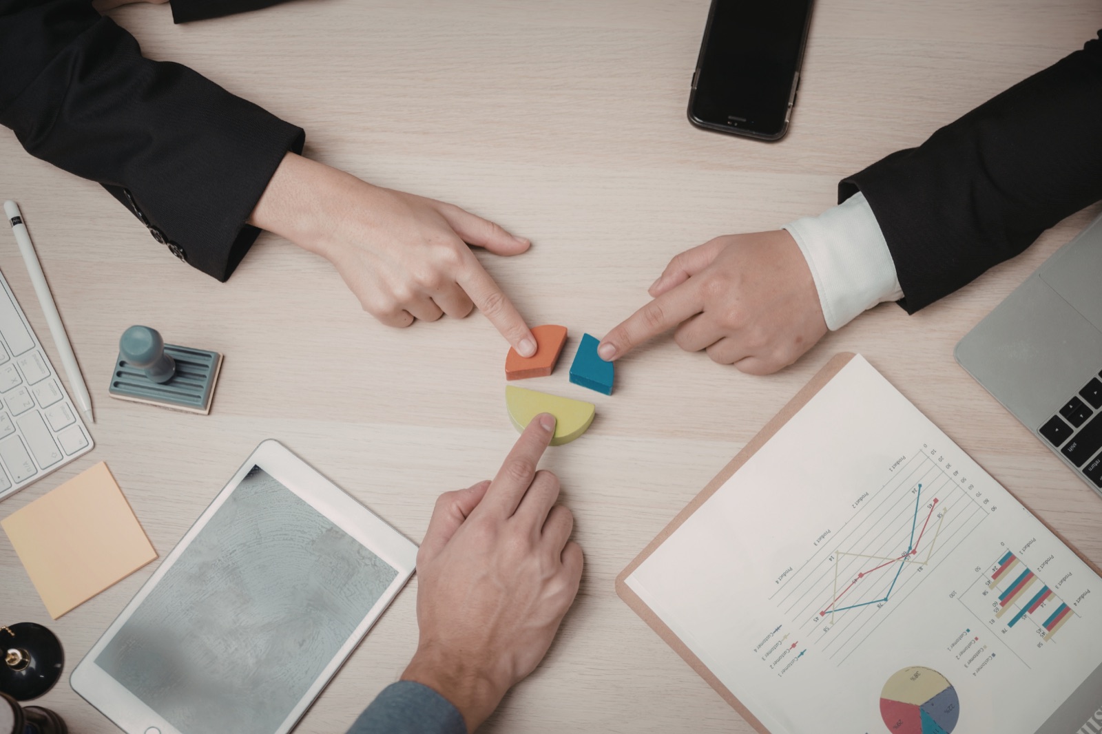 Hands assembling the colored pieces of a portfolio pie chart on a desk beside financial charts and devices